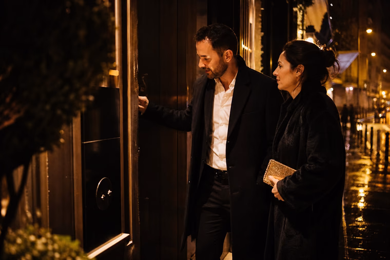 Un couple en tenue de soirée s’arrête devant l’entrée discrète d’un club, la nuit.