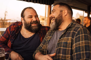 Deux hommes barbus et corpulents rient ensemble à la terrasse d’un bar de quartier.