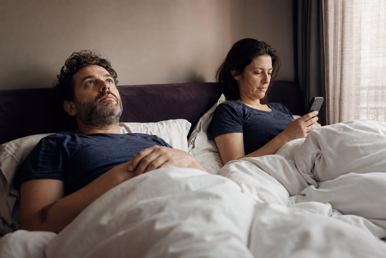 Un couple éveillé au lit le matin, chacun de son côté, dans une lumière douce.