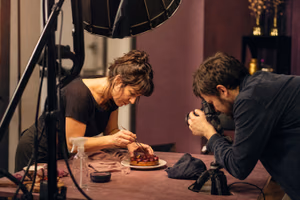 Une styliste culinaire retouche un dessert sur un plateau de studio, matériel d’éclairage visible.