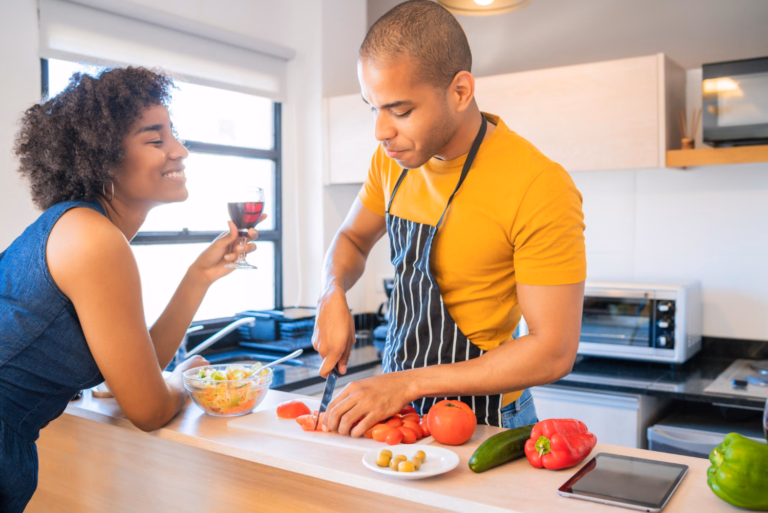 Un couple prépare un repas ensemble dans sa cuisine, un verre de vin posé sur le plan de travail.